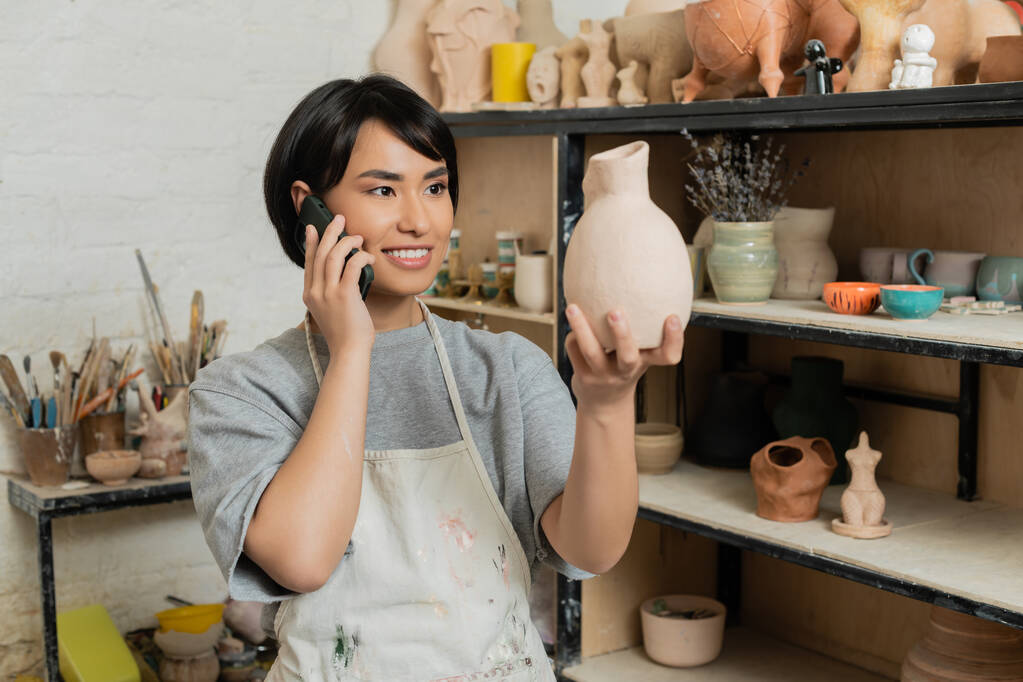 Jeune artisan asiatique souriant en tablier et vêtements de travail parlant sur smartphone et tenant une sculpture en céramique près d'un rack dans un studio d'art flou en arrière-plan, scène de studio de poterie - Photo, image
