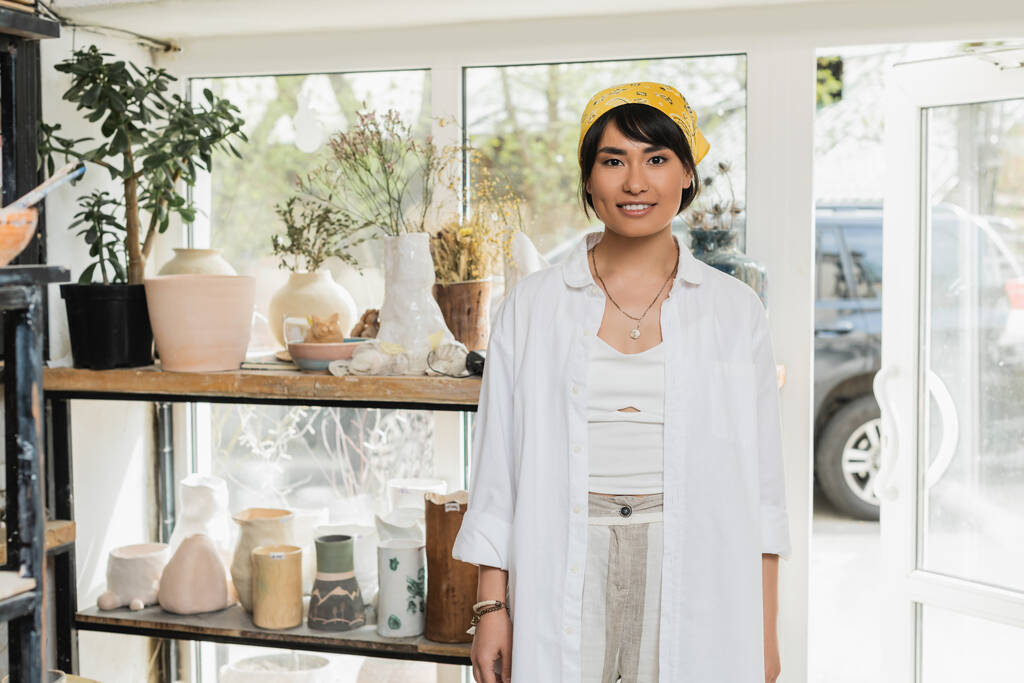 Retrato de sorrir jovem asiático feminino artesão em workwear e headscarf olhando para câmera enquanto está perto de esculturas cerâmicas no fundo em oficina de arte, cerâmica estúdio cena - Foto, Imagem