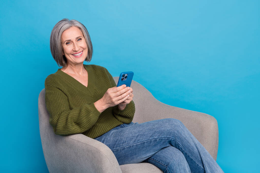 Ritratto di allegra donna positiva vestita elegante maglione kaki tenere smartphone leggere post in poltrona isolato su sfondo di colore blu. - Foto, immagini