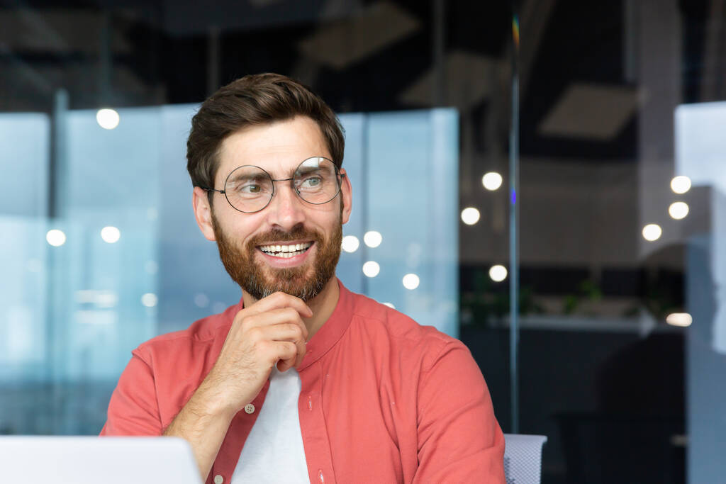 Улыбающийся мечтательный бизнесмен в офисе на рабочем месте, опытный и успешный программист-инженер улыбающийся и выглядывающий в окно, довольный результатами работы. - Фото, изображение