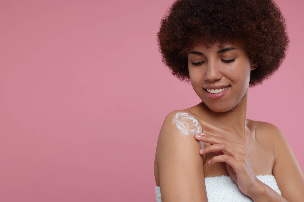 Hermosa mujer joven aplicando crema corporal en el hombro sobre fondo rosa, espacio para el texto - Foto, imagen