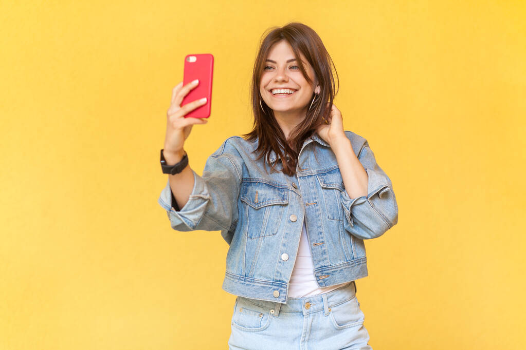 Muotokuva viehättävä kaunis hymyilevä nainen bloggaaja yllään denim takki tilalla matkapuhelin käsissä, joten selfie tai lähetykset livestream. Sisäuima studio ampui eristetty keltaisella pohjalla. - Valokuva, kuva