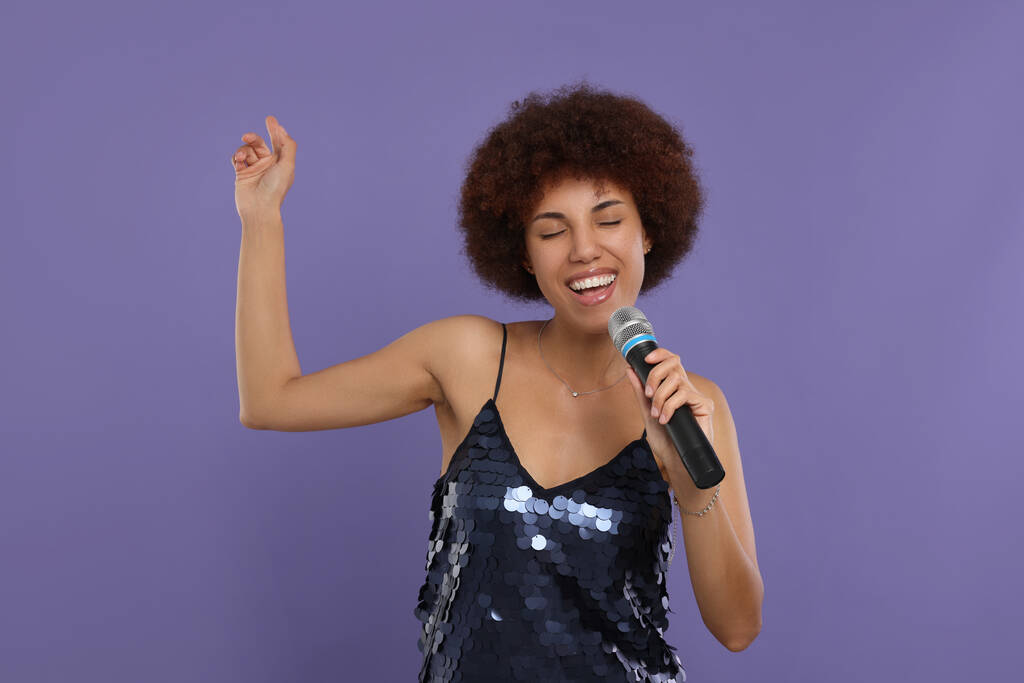 Σγουρή νεαρή γυναίκα με μικρόφωνο τραγουδά σε μωβ φόντο - Φωτογραφία, εικόνα