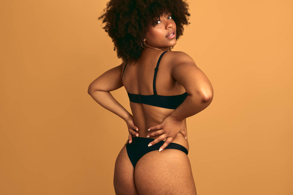 Jeune afro-américaine plus la taille femelle avec Afro coiffure portant lingerie noire debout avec les mains sur la taille en regardant la caméra sur fond orange - Photo, image