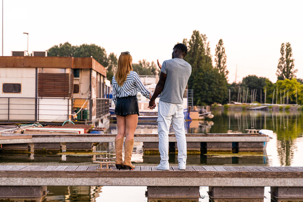 young multiracial loving couple holding hands on a quay of a river port at sunset - Photo, Image