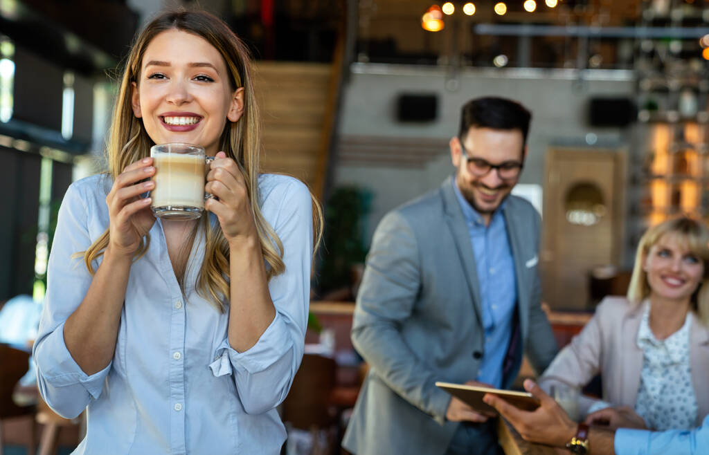 Porträt einer glücklichen jungen erfolgreichen Geschäftsfrau, die in einer Pause Kaffee trinkt. Im Hintergrund ihre Kollegen - Foto, Bild