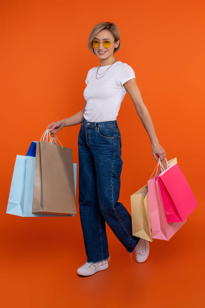 Радостная женщина в белой футболке и джинсах, стоящих с мешками для покупок изолированы на оранжевом фоне. - Фото, изображение