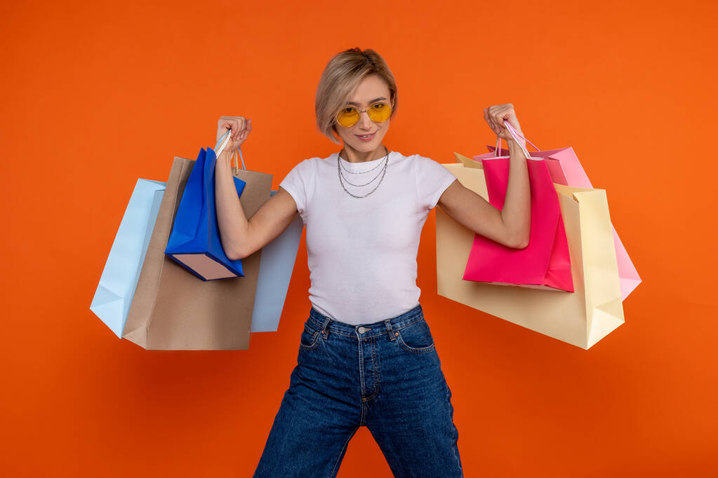 Beyaz tişörtlü ve kot pantolonlu komik güçlü kadın turuncu arka planda izole edilmiş alışveriş torbalarıyla kollarını kaldırdı.. - Fotoğraf, Görsel