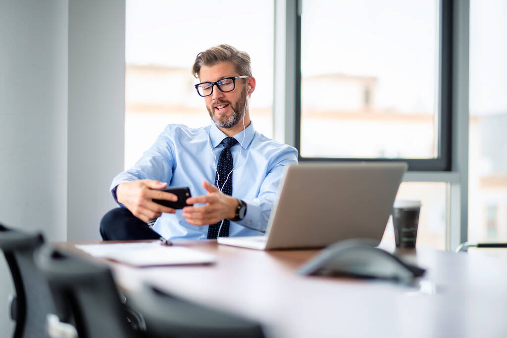 Portret van een zakenman die smartphone en oortjes gebruikt terwijl hij op kantoor zit en een videogesprek heeft. Professionele man draagt hemd en stropdas.  - Foto, afbeelding