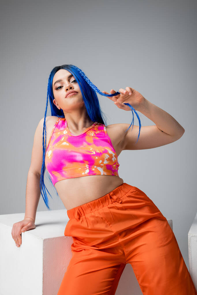 estilo rebelde, mujer joven tatuada con el pelo azul posando en ropa de colores cerca del cubo blanco sobre fondo gris, mirando a la cámara, individuo moderno, moda urbana, gen z - Foto, imagen