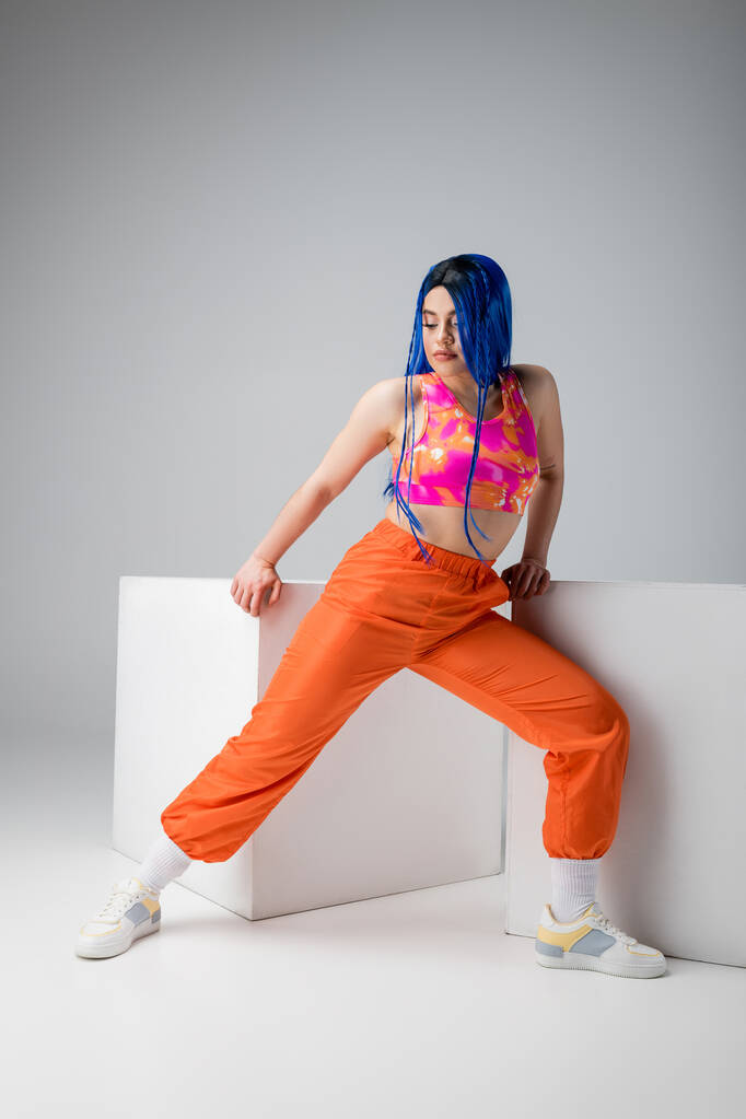 стильный вид, татуированная молодая женщина с голубыми волосами позирует в красочной одежде рядом с белыми кубиками на сером фоне, полная длина, индивидуализм, современный стиль, городская мода, яркий цвет, модель  - Фото, изображение