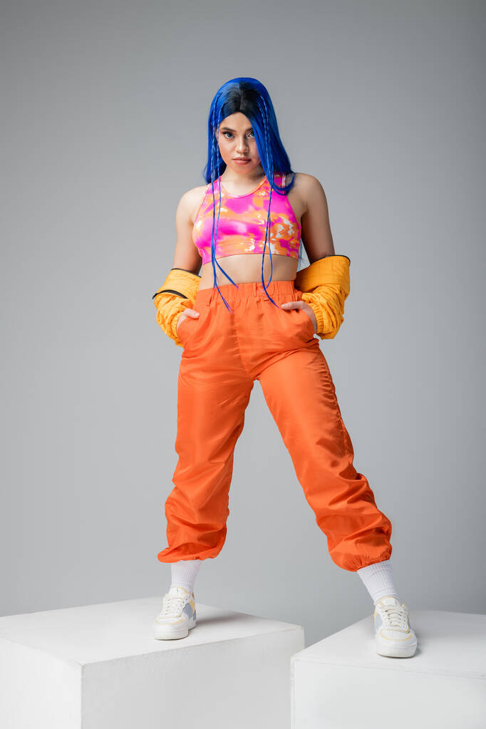 полная длина, мода вперед, татуированная женщина с голубыми волосами позирует с руками в карманах оранжевых брюк на сером фоне, стоя на белых кубиках, яркий цвет, женщина модель, городская мода  - Фото, изображение
