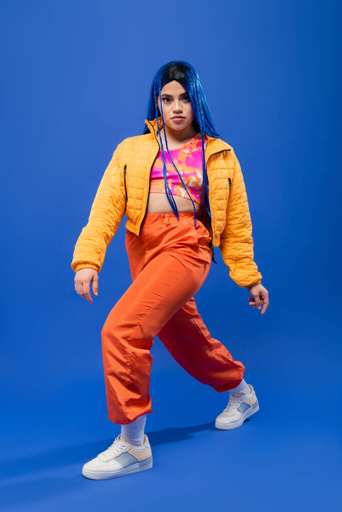 full length, fashion forward, νεαρή γυναίκα μοντέλο με μπλε μαλλιά ποζάρουν σε puffer σακάκι και πορτοκαλί παντελόνι σε μπλε φόντο, ζωντανό χρώμα, αστική μόδα, ατομικισμός - Φωτογραφία, εικόνα