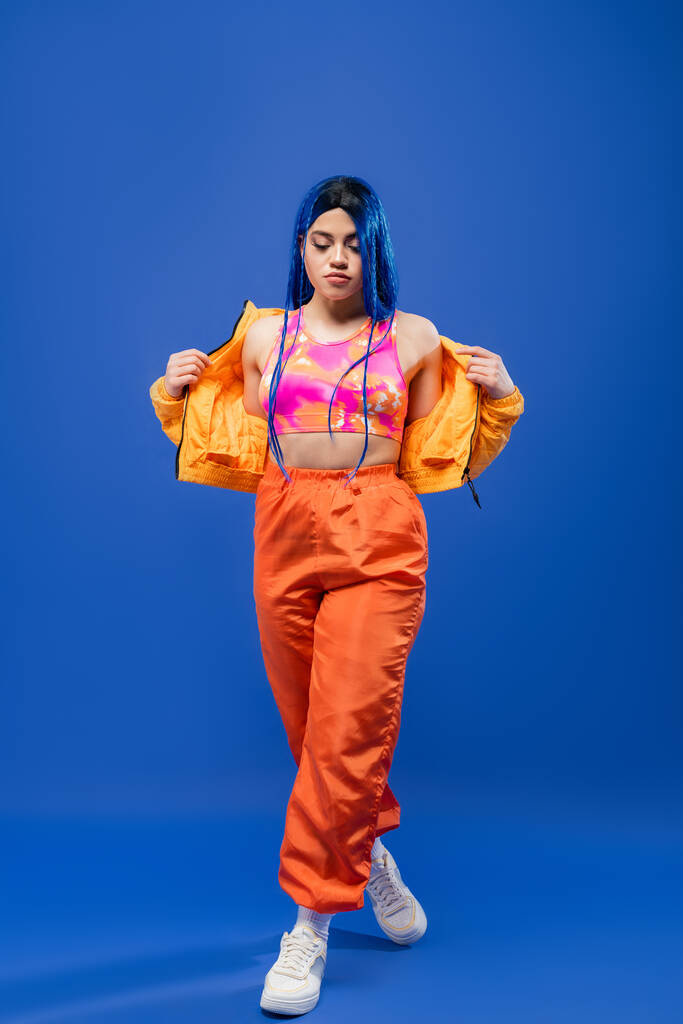 comprimento completo, cabelo tingido, estilo moderno, modelo feminino com cabelo azul posando em jaqueta e calças laranja no fundo azul, cor vibrante, moda urbana, individualismo, jovem  - Foto, Imagem