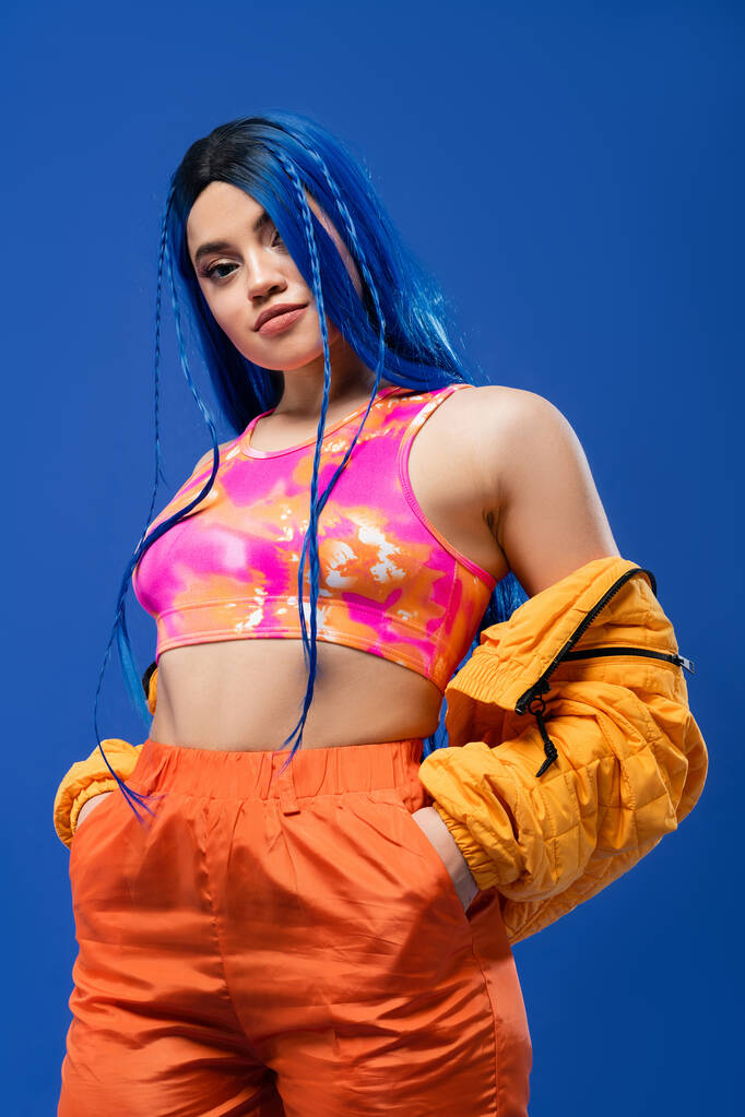 colorful clothes, dyed hair, female model with blue hair posing with hands in pockets on blue background, puffer jacket, vibrant color, urban fashion, individualism, young woman with funky look  - Photo, Image