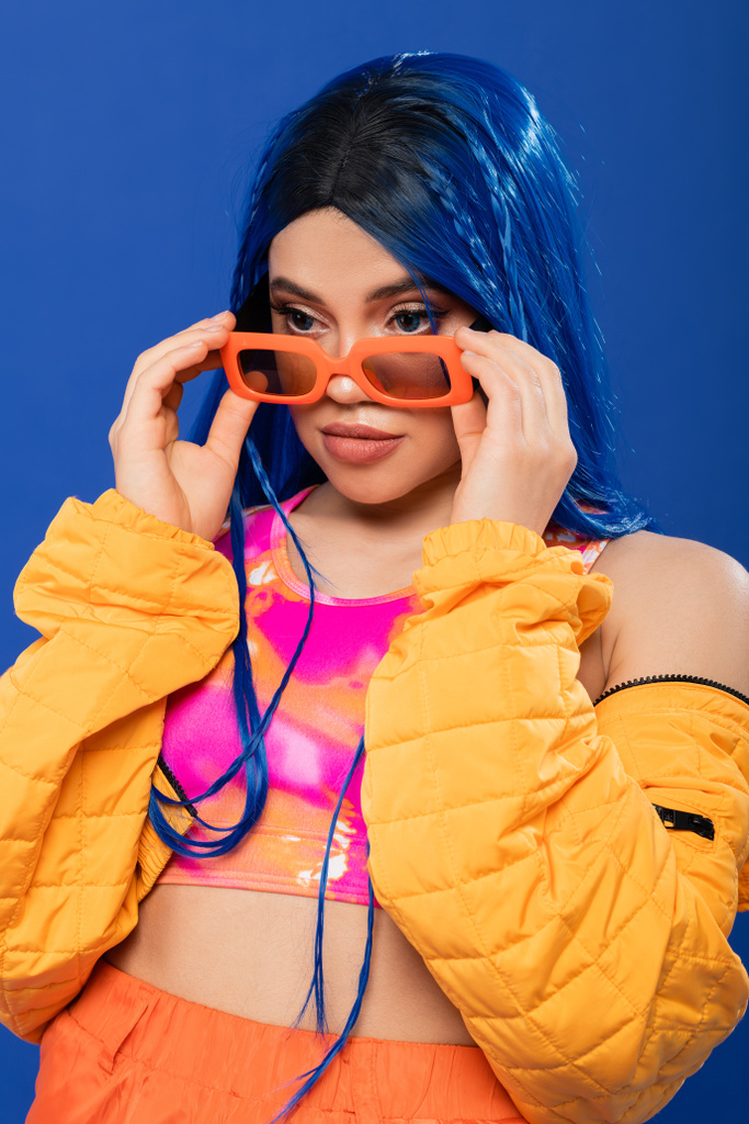 moda y estilo, modelo femenino joven con pelo azul y trenzas con gafas de sol de color naranja aisladas sobre fondo azul, generación z, estilo rebelde, ropa colorida, individualismo, mujer moderna  - Foto, imagen