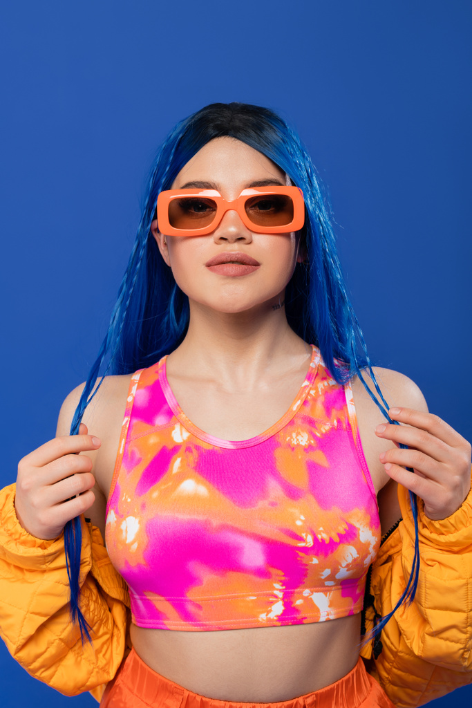 δήλωση μόδας, νεαρή γυναίκα μοντέλο με μπλε μαλλιά και πλεξούδες θέτουν σε μοντέρνα γυαλιά ηλίου που απομονώνονται σε μπλε φόντο, γενιά z, επαναστατικό στυλ, πολύχρωμα ρούχα, ατομικισμός, σύγχρονη γυναίκα  - Φωτογραφία, εικόνα