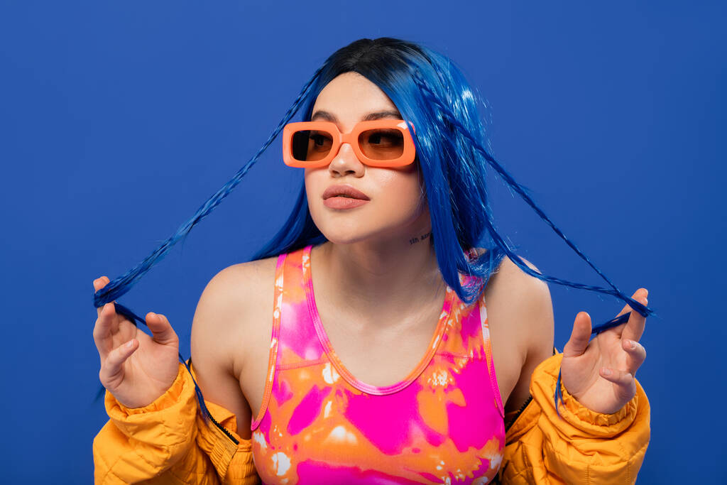 fashion statement, young female model with blue hair touching braids and trendy sunglasses isolated on blue background, generation z, rebel style, colorful clothes, individualism, modern woman  - Photo, Image