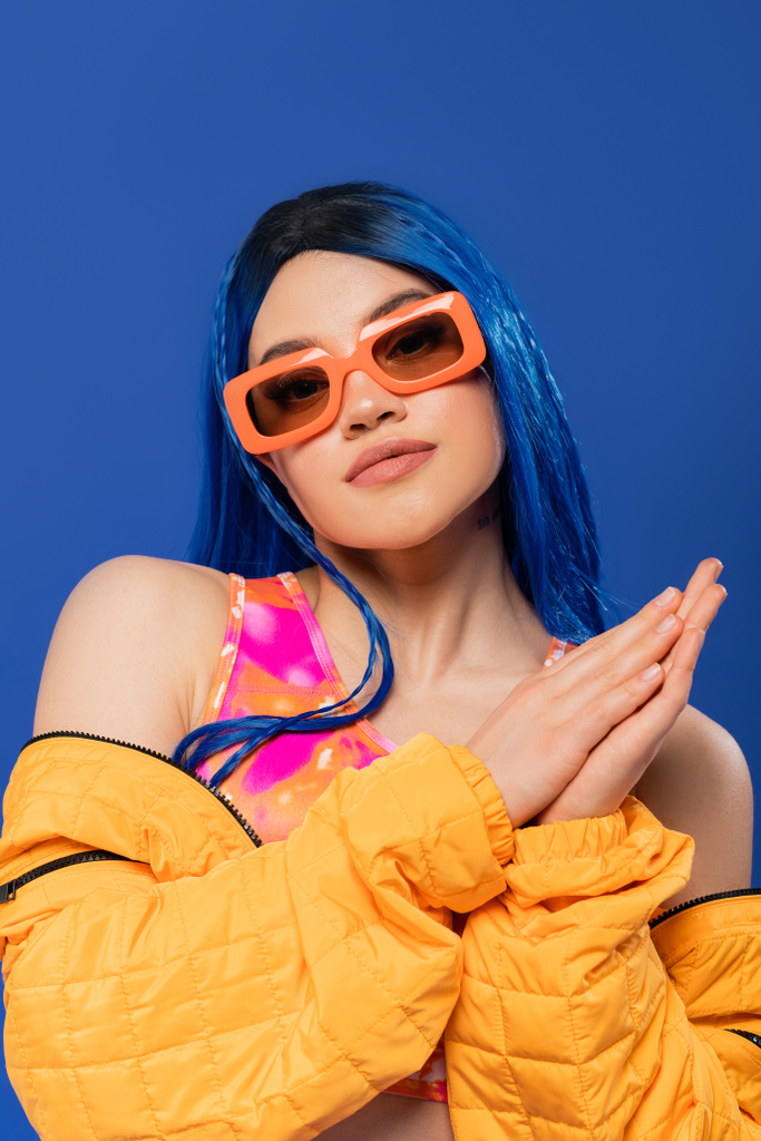 δήλωση μόδας, νεαρή γυναίκα μοντέλο με μπλε μαλλιά και μοντέρνα γυαλιά ηλίου που απομονώνονται σε μπλε φόντο, γενιά z, επαναστατικό στυλ, πολύχρωμα ρούχα, ατομικισμός, σύγχρονη γυναίκα κοιτάζοντας κάμερα  - Φωτογραφία, εικόνα