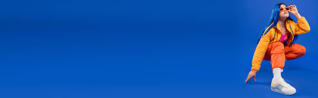 generation z, full length of pretty female model with blue hair and trendy glasses sitting on hunches on blue background, επαναστατικό στυλ, μοντέρνα μόδα, μοντέρνο αξεσουάρ, banner - Φωτογραφία, εικόνα