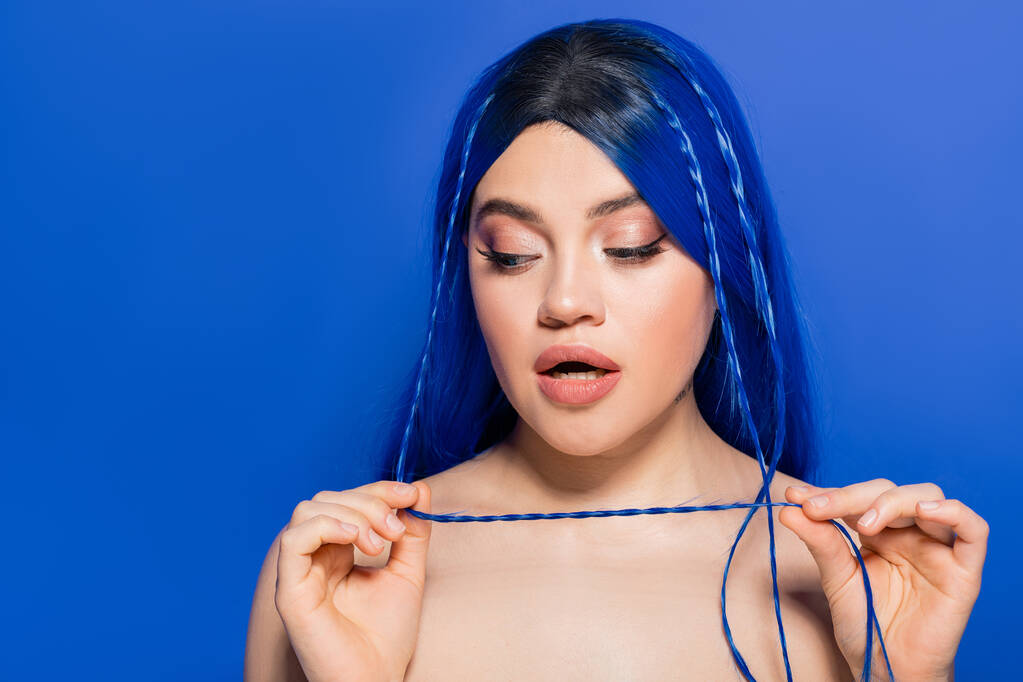 lebendige Jugend, Selbstausdruck, emotional, Porträt einer überraschten jungen Frau mit gefärbten Haaren, die auf blauem Hintergrund posiert, Haarfarbe, Individualismus, weibliches Modell mit Make-up und trendiger Frisur  - Foto, Bild