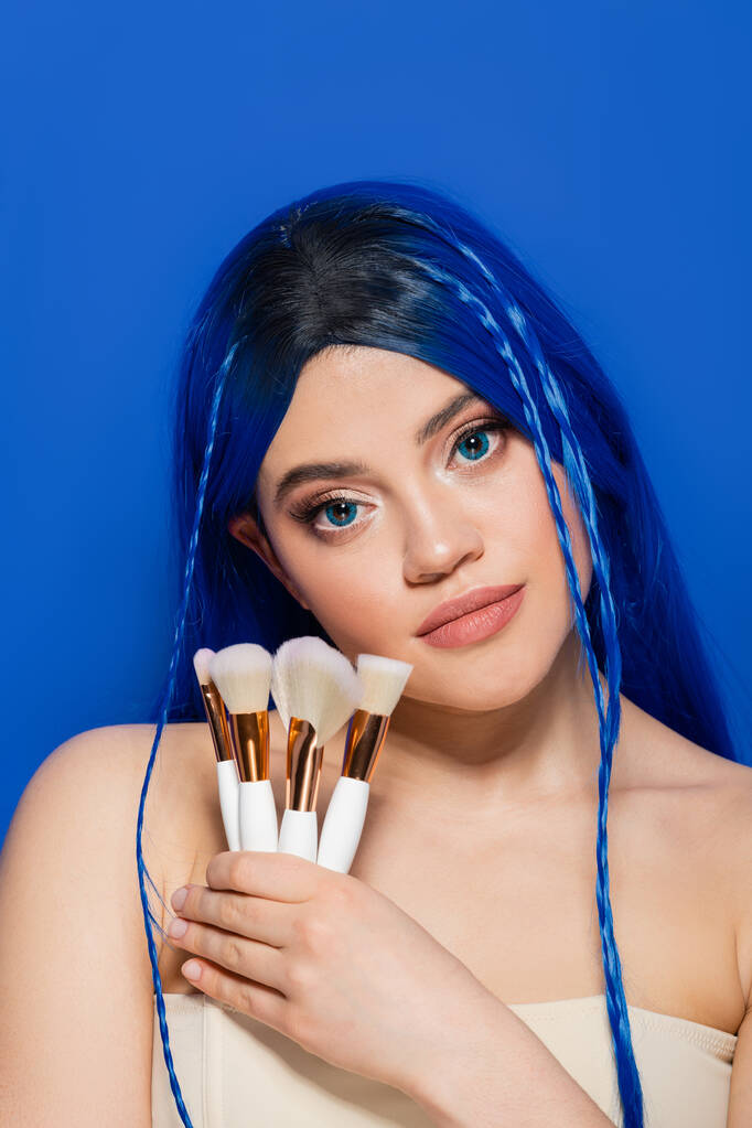 beauty industry, individualism, young woman with vibrant hair and eyes looking at camera while holding cosmetic brushes on blue background, makeup, beauty trends, visage, youth, self expression  - Photo, Image