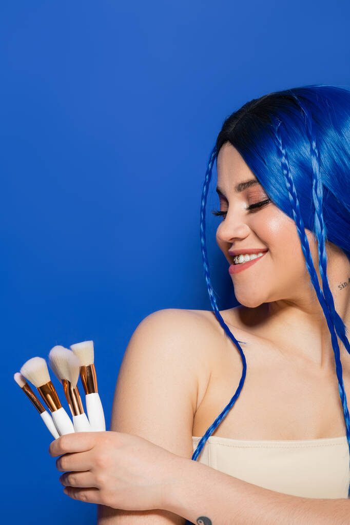 beauty industry, individualism, joyful young woman with vibrant hair and eyes holding cosmetic brushes on blue background, makeup, beauty trends, visage, youth, self expression  - Photo, Image