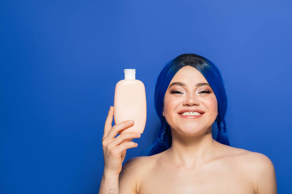 Güzellik trendleri, saç bakımı konsepti, parlak saçlı mutlu bir kadının portresi mavi arka planda çıplak omuzlarıyla poz vermesi, şampuanlı kozmetik bir şişe tutması, reklam.  - Fotoğraf, Görsel