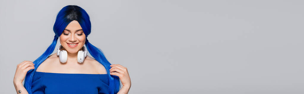 Musikliebhaberin, glückliche junge Frau mit blauen Haaren und kabellosen Kopfhörern, die auf grauem Hintergrund lächelt, lebendige Jugend, Individualismus, moderne Subkultur, Selbstausdruck, Tätowierung, Sound, Banner - Foto, Bild