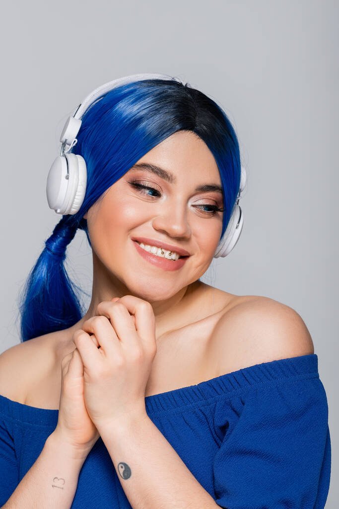 αυτοέκφραση, χαρούμενη νεαρή γυναίκα με μπλε μαλλιά ακούει μουσική σε ασύρματα ακουστικά σε γκρι φόντο, ζωντανή νεολαία, ατομικισμός, σύγχρονη υποκουλτούρα, τατουάζ, ήχο  - Φωτογραφία, εικόνα