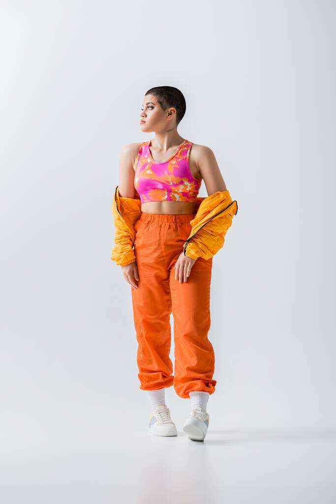 generation z, fashionable young woman with short hair posing in puffer jacket and vibrant pants on grey background, looking away, outerwear, modern subculture, youthful energy, full length  - Foto, immagini