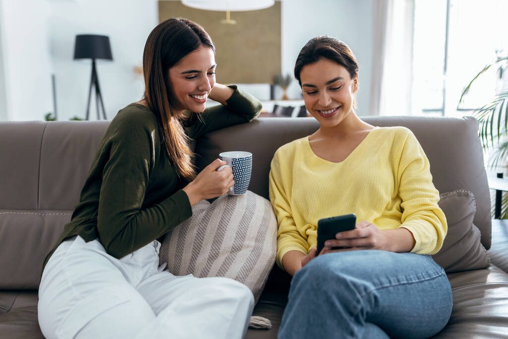 Съемка двух улыбающихся молодых женщин разговаривающих во время просмотра смартфона сидя на диване в гостиной дома. - Фото, изображение