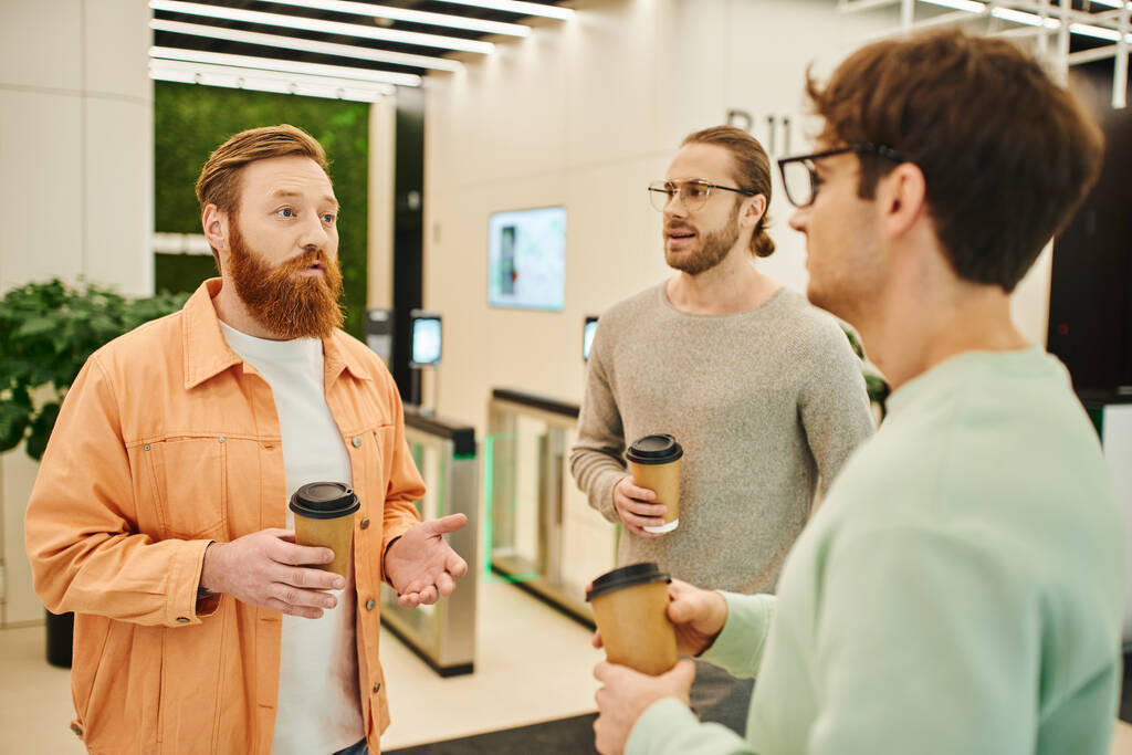 poważny mężczyzna z brodą rozmawiający z kolegami w okularach trzymającymi papierowe kubki z napojami na wynos podczas przerwy kawowej we współczesnym biurze współpracowników, partnerstwo i współpraca w nowoczesnym biznesie - Zdjęcie, obraz