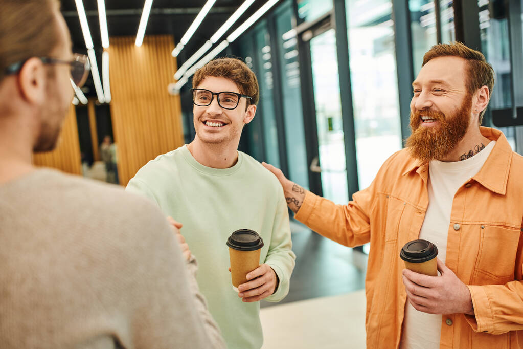 позитивний бородатий чоловік торкається плеча щасливого колеги, тремтячи руками з бізнесменом на розмитому фоні, успішні підприємці з кавою йдуть підтверджуючи угоду в сучасному офісі - Фото, зображення