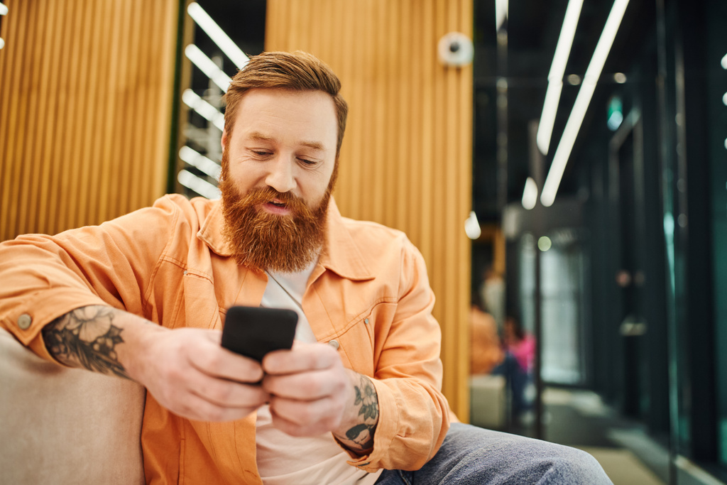 бородатый и татуированный бизнесмен в стильной повседневной одежде, работающий на мобильном телефоне, сидя на удобном диване в гостиной современного коворкинга на размытом фоне - Фото, изображение