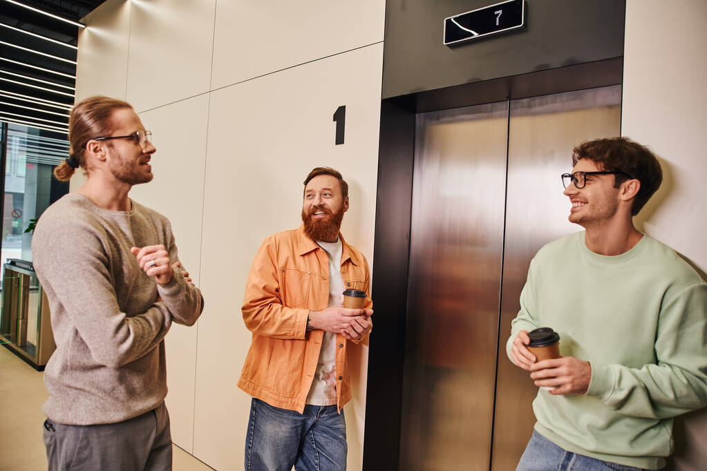zadowoleni współpracownicy biznesowi w stylowych ubraniach casual trzymając drinki na wynos w kubkach papierowych czekając na windę, odnoszący sukcesy przedsiębiorcy rozmawiający podczas przerwy kawowej w nowoczesnym biurze - Zdjęcie, obraz