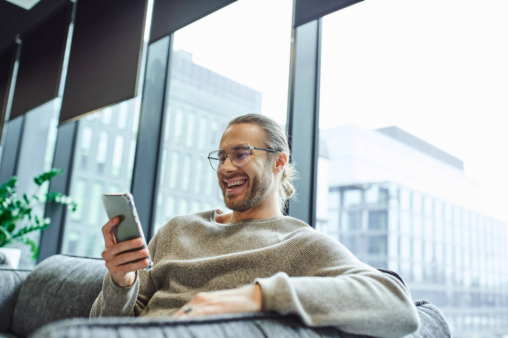 excited and stylish businessman in eyeglasses and casual clothes sitting in contemporary office lounge near large windows and looking at mobile phone, business inspiration and positivity - Photo, Image