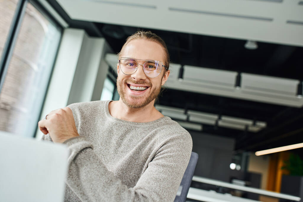 профессиональный снимок восторженного бизнесмена в стильной одежде и очках, улыбающегося в камеру возле ноутбука на размытом переднем плане в современном офисе, бизнес-продуктивности и концепции успеха - Фото, изображение