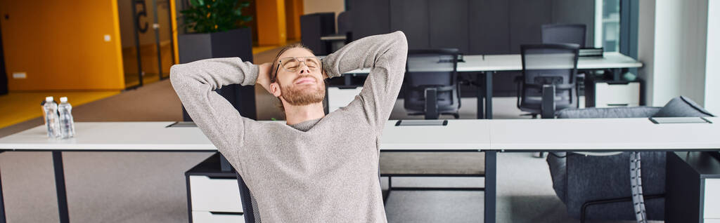 κουρασμένος επιχειρηματίας με κομψά γυαλιά ηλίου και casual ρούχα κάθεται με κλειστά μάτια και τα χέρια πίσω από το κεφάλι, ενώ στηρίζεται στο χώρο εργασίας στο σύγχρονο περιβάλλον γραφείου, πανό - Φωτογραφία, εικόνα