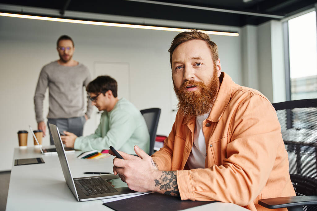 bearded and tattooed businessman in stylish casual clothes holding smartphone and looking at camera near laptop and colleagues working on blurred background in contemporary coworking space - Photo, Image