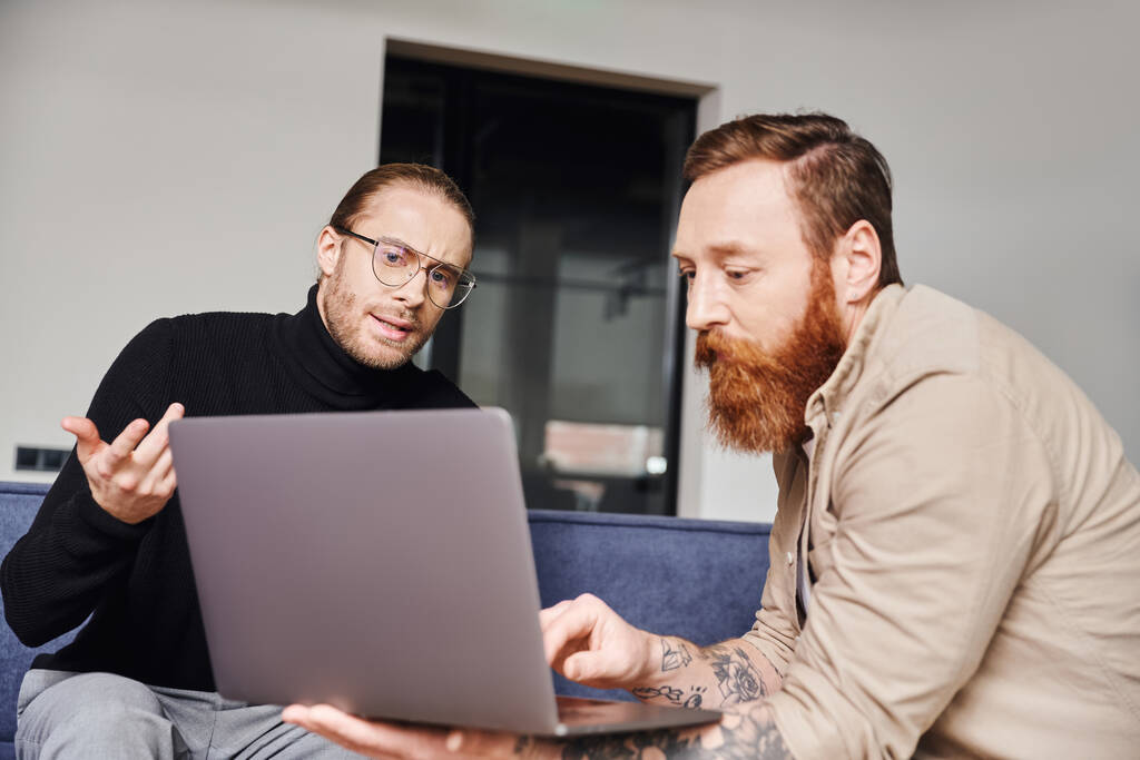 biznesmen w czarnym golfie i okularach gestykulujący podczas rozmowy z brodatym, wytatuowanym przedsiębiorcą pokazującym projekt startupowy na laptopie w nowoczesnym biurze, koncepcja partnerstwa biznesowego - Zdjęcie, obraz