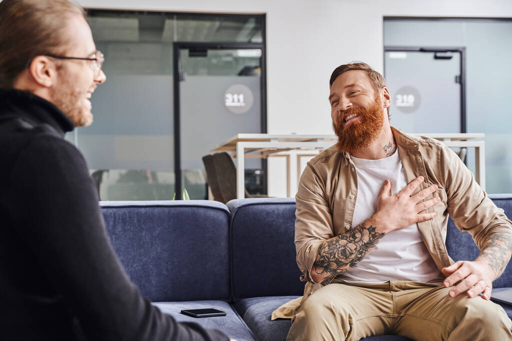 dankbare, bebaarde en getatoeëerde ondernemer raakt borst tijdens gesprek met zakenpartner op bank in moderne kantooromgeving, business collaboration concept - Foto, afbeelding