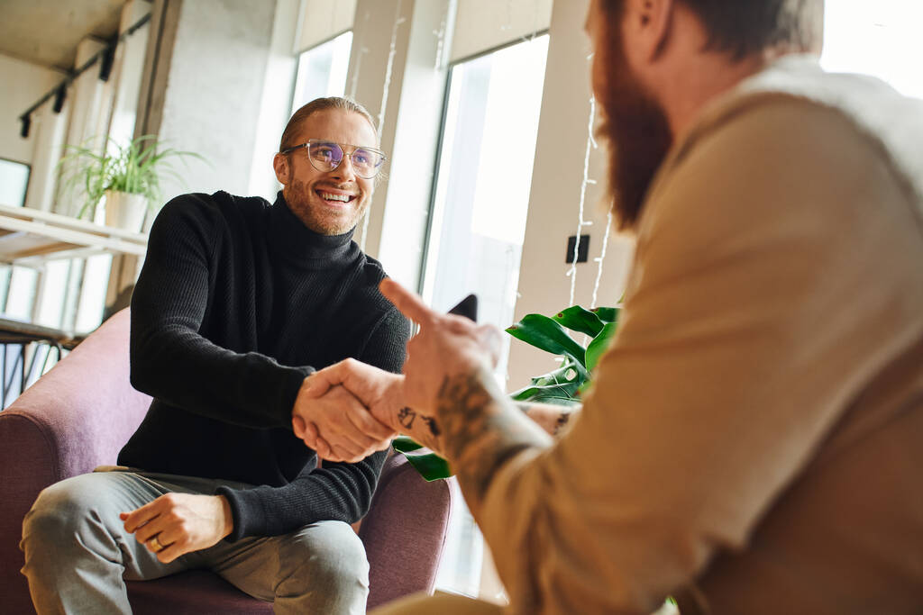 happy entrepreneur in eyeglasses and black turtleneck shaking hands and closing deal with tattooed businessman pointing with finger on blurred foreground, successful partnership concept - Photo, Image