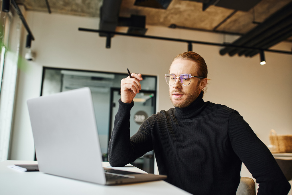thoughtful entrepreneur in black turtleneck and eyeglasses holding pen and looking at laptop while working on startup planning in contemporary coworking office, business lifestyle concept - Photo, Image
