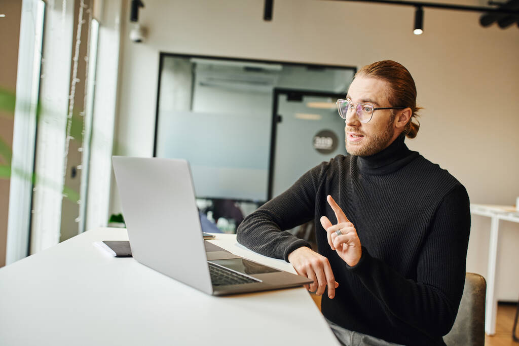 stylish businessman in black turtleneck and eyeglasses showing attention gesture and talking during video chat on laptop at modern workplace in coworking office, business lifestyle concept - Photo, Image