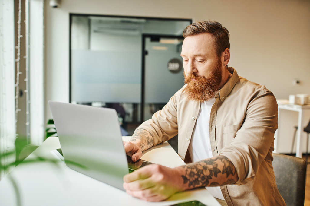 serious bearded and tattooed entrepreneur working on startup project while sitting near laptop at workplace in modern coworking environment, business lifestyle concept - Photo, Image