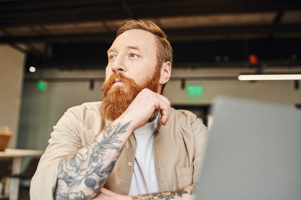 portrait of pensive, tattooed and bearded entrepreneur sitting at workplace, looking away and thinking near laptop on blurred foreground in modern office, business lifestyle concept - Photo, Image