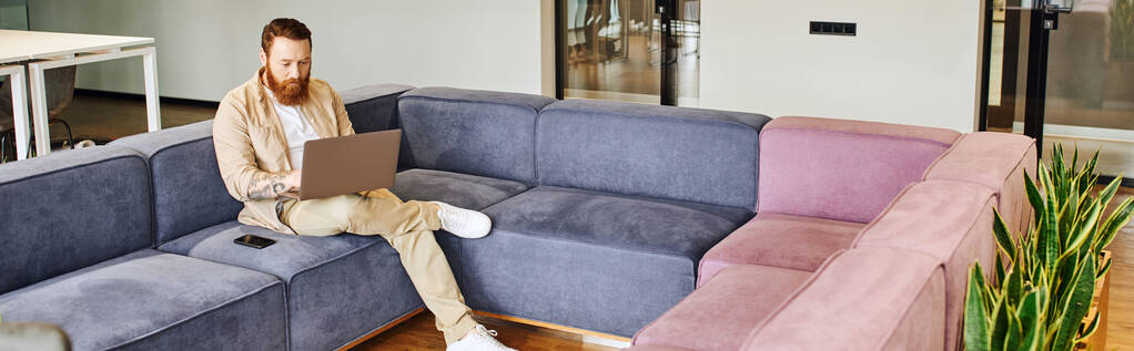 volle Länge der stilvollen, bärtigen und tätowierten Unternehmer sitzt auf bequemen Sofa in der Nähe von Smartphone und arbeitet am Computer in der Lounge des zeitgenössischen Büros, Business-Lifestyle-Konzept, Banner - Foto, Bild