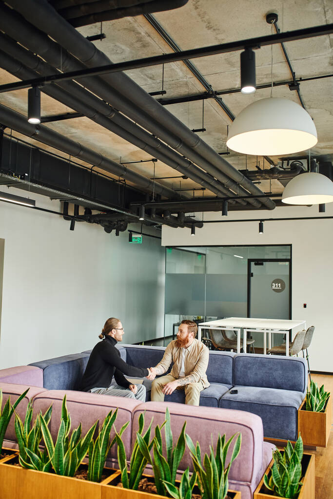 business partners in stylish casual clothes shaking hands and confirming agreement while sitting on cozy couch in modern office lounge with high tech interior, partnership and success concept - Photo, Image