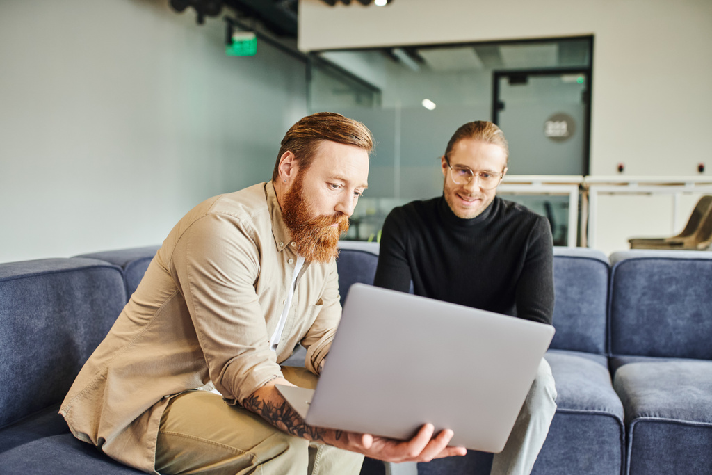 bearded and tattooed entrepreneur showing laptop with business project to pleased colleague in lounge of contemporary office environment, business partnership concept - Photo, Image